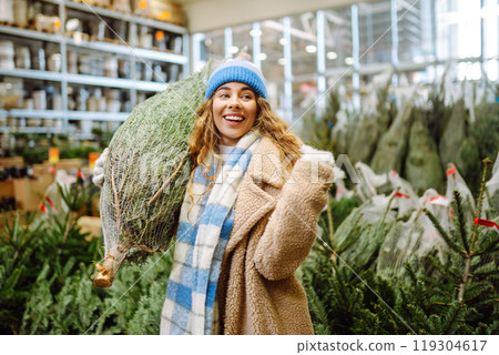young smiling woman chooses Christmas tree to buy. Happy woman buying New Year tree for Christmas. young smiling woman chooses Christmas tree to buy. Happy woman buying New Year tree for Christmas. 119304617