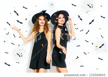 portrait of two happy young women in black witch halloween costumes. firecrackers in the background. 119304643
