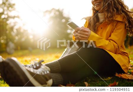 A smiling woman sits outdoors on the lawn among yellow autumn leaves with a phone in her hands. 119304931