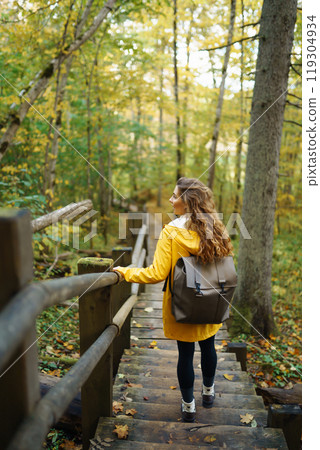 Woman in a yellow coat with backpack walks along wooden staircase, enjoying autumn scenery in forest 119304934