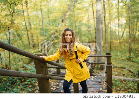 Woman in a yellow coat with backpack walks along wooden staircase, enjoying autumn scenery in forest 119304935