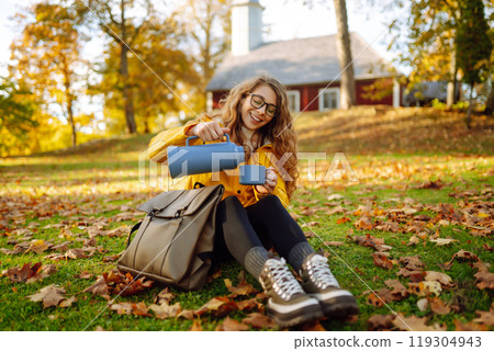 Woman traveler in yellow coat sits in clearing among yellow leaves and drinks hot drink from thermos Woman traveler in yellow coat sits in clearing among yellow leaves and drinks hot drink from thermos 119304943