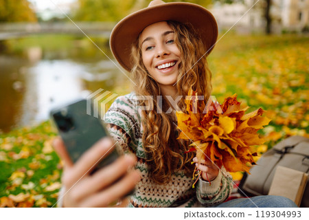 Selfie time. Beautiful woman takes selfie on smartphone in hat on meadow in autumn park on plaid. Selfie time. Beautiful woman takes selfie on smartphone in hat on meadow in autumn park on plaid. 119304993