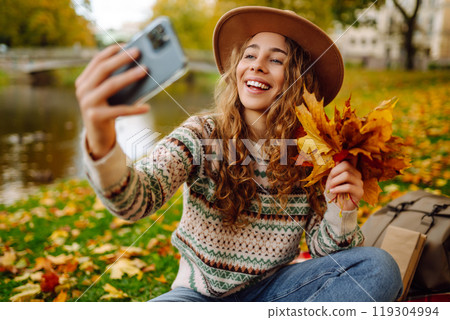 Selfie time. Beautiful woman takes selfie on smartphone in hat on meadow in autumn park on plaid. Selfie time. Beautiful woman takes selfie on smartphone in hat on meadow in autumn park on plaid. 119304994
