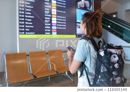 The girl goes on a flight in the airport, holds a passport and tickets in her hands, looks at the gate number in search of her plane, the woman goes on a trip with a backpack. 119305149