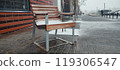 A snow-covered bench sits alone on a city sidewalk during a winter storm in an urban area 119306547