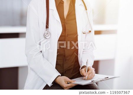 Unknown woman-doctor writing something at clipboard while sitting in sunny clinic, close-up. Medicine concept Unknown woman-doctor writing something at clipboard while sitting in sunny clinic, close-up. Medicine concept 119307367