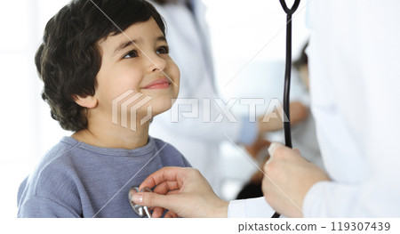 Doctor-woman examining a child patient by stethoscope. Cute arab boy and his brother at physician appointment. Medicine concept Doctor-woman examining a child patient by stethoscope. Cute arab boy and his brother at physician appointment. Medicine concept 119307439