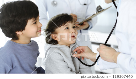 Woman-doctor examining a child patient by stethoscope. Cute arab toddler and his brother at physician appointment. Medicine concept Woman-doctor examining a child patient by stethoscope. Cute arab toddler and his brother at physician appointment. Medicine concept 119307440