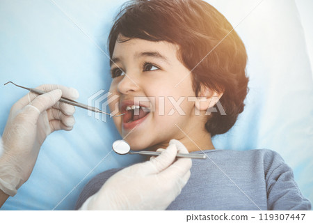 Cute arab boy sitting at dental chair with open mouth during oral checking up with doctor. Visiting sunny dentist office 119307447
