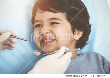Cute arab boy sitting at dental chair with open mouth during oral checking up with doctor. Visiting sunny dentist office 119307448