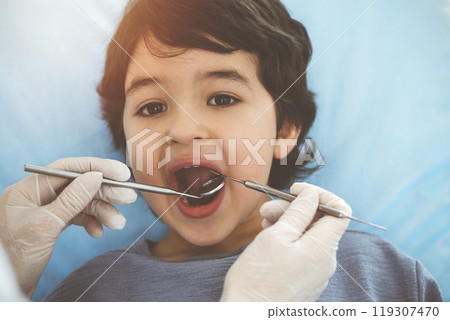 Cute arab boy sitting at dental chair with open mouth during oral checking up with doctor. Visiting sunny dentist office Cute arab boy sitting at dental chair with open mouth during oral checking up with doctor. Visiting sunny dentist office 119307470