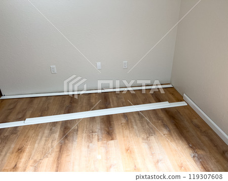 A room with fresh wooden flooring, two windows, and white vinyl baseboards positioned along the walls, ready for installation. The space is bright and shows progress in the renovation process. A room with fresh wooden flooring, two windows, and white vinyl baseboards positioned along the walls, ready for installation. The space is bright and shows progress in the renovation process. 119307608