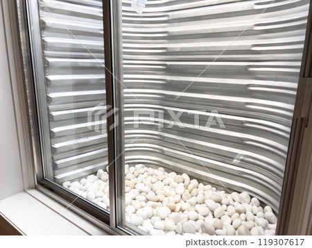 A clear view from a basement window showing a corrugated metal window well lined with smooth white decorative stones at the base. The metal surface appears freshly installed and clean. A clear view from a basement window showing a corrugated metal window well lined with smooth white decorative stones at the base. The metal surface appears freshly installed and clean. 119307617