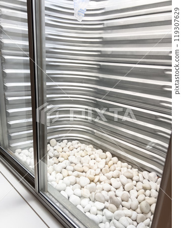 A clear view from a basement window showing a corrugated metal window well lined with smooth white decorative stones at the base. The metal surface appears freshly installed and clean. A clear view from a basement window showing a corrugated metal window well lined with smooth white decorative stones at the base. The metal surface appears freshly installed and clean. 119307629