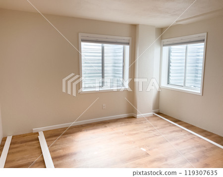 A room with fresh wooden flooring, two windows, and white vinyl baseboards positioned along the walls, ready for installation. The space is bright and shows progress in the renovation process. 119307635