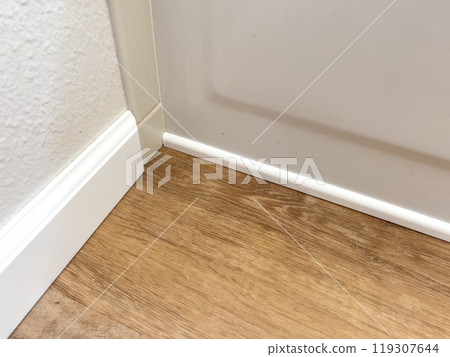White vinyl baseboards and trim pieces are neatly stacked against a wall, prepared for installation in a room with wooden flooring. The renovation project is in progress, ready to complete the room 119307644
