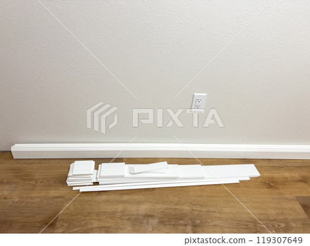 White vinyl baseboards and trim pieces are neatly stacked against a wall, prepared for installation in a room with wooden flooring. The renovation project is in progress, ready to complete the room 119307649