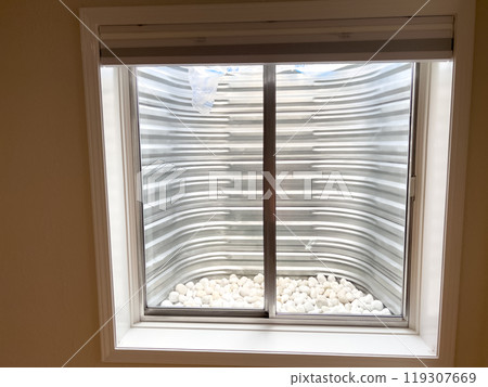 A clear view from a basement window showing a corrugated metal window well lined with smooth white decorative stones at the base. The metal surface appears freshly installed and clean. 119307669