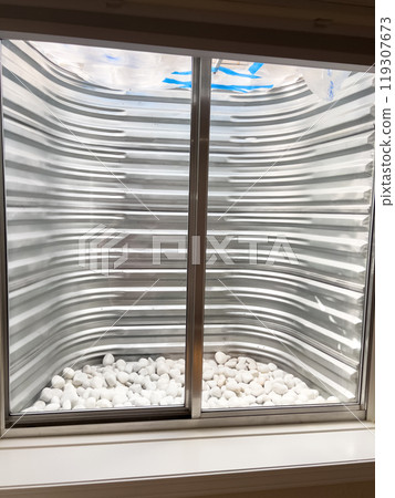 A clear view from a basement window showing a corrugated metal window well lined with smooth white decorative stones at the base. The metal surface appears freshly installed and clean. 119307673