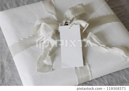 Closeup of gift box with empty paper sale,gift tag mockup, white silk ribbon on linen tablecloth. Christmas, wedding or birthday present. Selective focus, blurred background. Gift wrapping concept. 119308571
