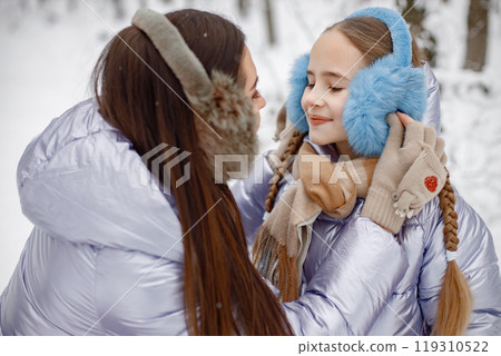 Mother with her child having fun in forest on winter day. Girls wearing purple warm jackets and earmuffs. Female models posing for a photo. Mother with her child having fun in forest on winter day. Girls wearing purple warm jackets and earmuffs. Female models posing for a photo. 119310522