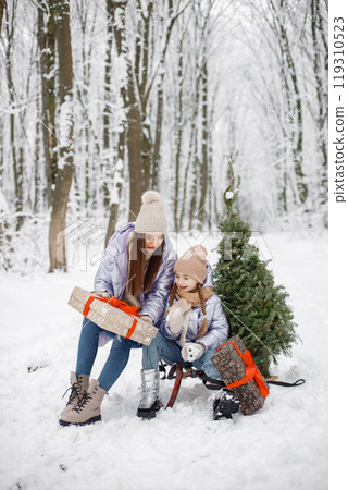 Mother with her child having fun in forest on snow day. Girls wearing purple warm jackets and knitted hats. Girls holding a gift boxes and little Christmas tree. Mother with her child having fun in forest on snow day. Girls wearing purple warm jackets and knitted hats. Girls holding a gift boxes and little Christmas tree. 119310523