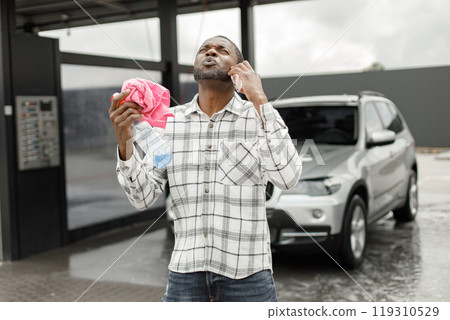 Man washing luxury car on a car wash using sprayer bottle and a rag. Black man talking on the phone and holding in his hands rose rag and sprayer bottle for polishing his car. Man wearing plaid shirt 119310529