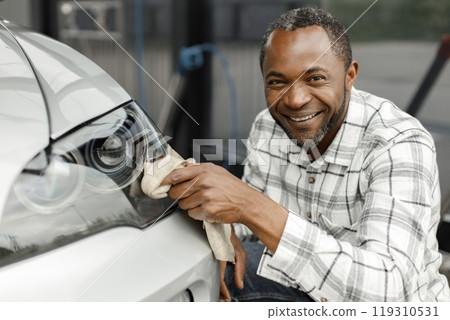 Man washing luxury car on a car wash with a rag. Black man wiping his car. Man wearing plaid shirt, smiling and looking at camera. Man washing luxury car on a car wash with a rag. Black man wiping his car. Man wearing plaid shirt, smiling and looking at camera. 119310531