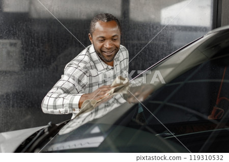 Man washing luxury car on a car wash with a rag. Black man wiping his car. Man wearing plaid shirt, smiling. Man washing luxury car on a car wash with a rag. Black man wiping his car. Man wearing plaid shirt, smiling. 119310532