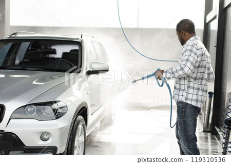 Man washing luxury car on a car wash with a pressure washer. Black man using high pressure hose with water. Man wearing plaid shirt. 119310536