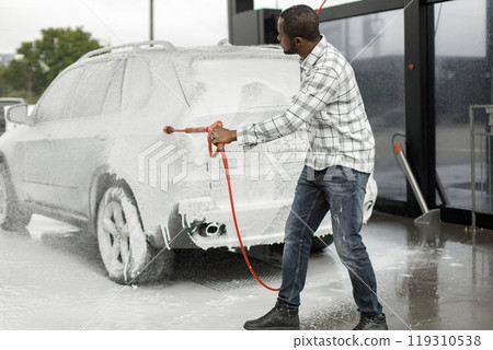 Man washing luxury car on a car wash with a pressure washer. Black man using high pressure hose with foam. Man wearing plaid shirt. Man washing luxury car on a car wash with a pressure washer. Black man using high pressure hose with foam. Man wearing plaid shirt. 119310538