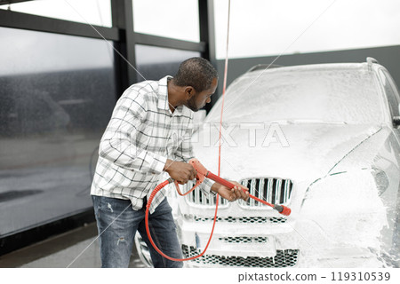 Man washing luxury car on a car wash with a pressure washer. Black man using high pressure hose with soap. Man wearing plaid shirt. Man washing luxury car on a car wash with a pressure washer. Black man using high pressure hose with soap. Man wearing plaid shirt. 119310539
