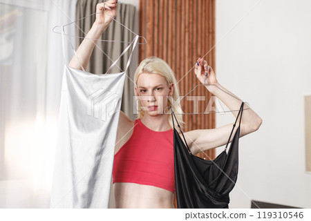 Man with make up on his face standing in a bedroom in his flat. Man wearing fashionable pink top, white trousers. Man choosing what to wear: black or white dress. Man with make up on his face standing in a bedroom in his flat. Man wearing fashionable pink top, white trousers. Man choosing what to wear: black or white dress. 119310546