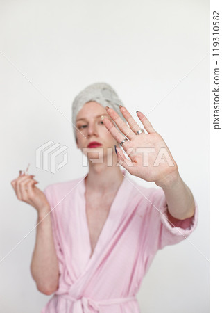 Young man with make up on his face doing manicure. Man wearing pink dressing gown and has a towel on his head. Man isolated on white background. Young man with make up on his face doing manicure. Man wearing pink dressing gown and has a towel on his head. Man isolated on white background. 119310582