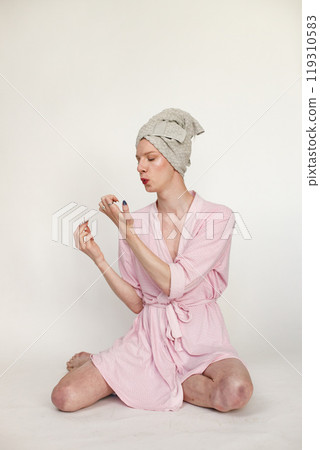 Young man with make up on his face doing manicure. Man wearing pink dressing gown and has a towel on his head. Man isolated on white background. 119310583