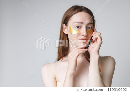 Close up beauty portrait of an attractive young woman applying gel patches on her face isolated over white background. Woman has no shirt. Woman has long red hair. 119310602