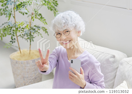 Senior woman making a peace sign with a smartphone in the living room Senior woman making a peace sign with a smartphone in the living room 119313032