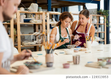 Female teacher and mentor helps students learn how to make patterns on ceramic plates using paint and brush in pottery workshop Female teacher and mentor helps students learn how to make patterns on ceramic plates using paint and brush in pottery workshop 119313953