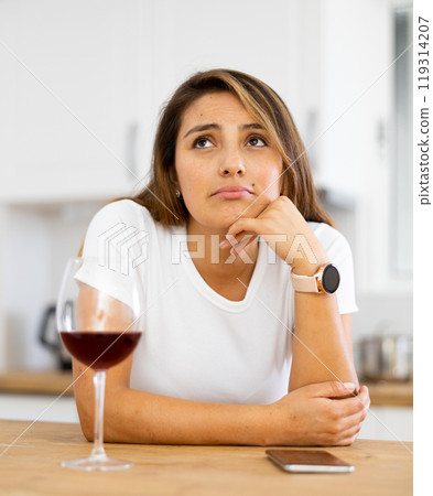 Frustrated young Hispanic woman with phone drinking wine in home kitchen Frustrated young Hispanic woman with phone drinking wine in home kitchen 119314207