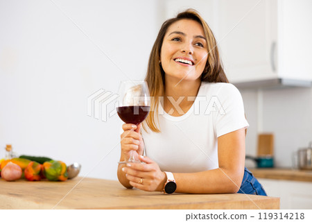 Cheerful young Latina drinking wine in home kitchen 119314218