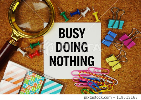 Busy Doing Nothing text on paper card with magnifying glass and stationery on cork board background Busy Doing Nothing text on paper card with magnifying glass and stationery on cork board background 119315165