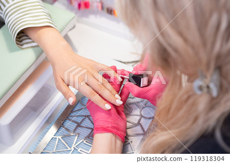 Nail technician in pink gloves skillfully applies nude gel polish to a clients hand while ensuring perfect nails. 119318304