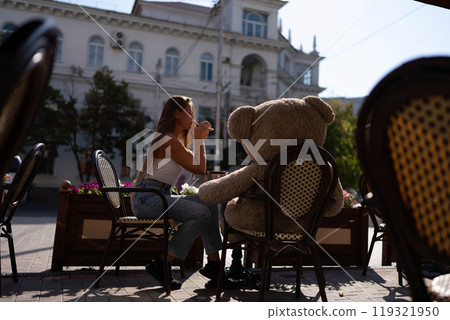 A woman sits cafe with a teddy bear next to her. The scene is set in a city with several chairs and tables around her. The woman is enjoying her time at the outdoor cafe. A woman sits cafe with a teddy bear next to her. The scene is set in a city with several chairs and tables around her. The woman is enjoying her time at the outdoor cafe. 119321950