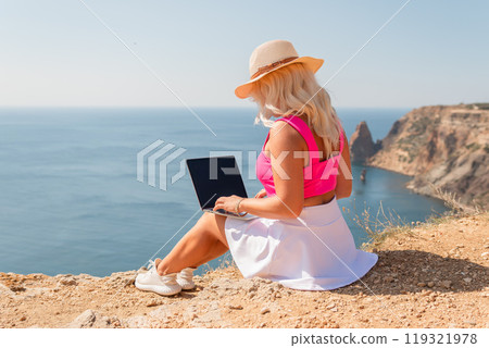 A woman is sitting on a rocky beach with a laptop open in front of her. She is wearing a pink top and a straw hat. Concept of relaxation and leisure. 119321978