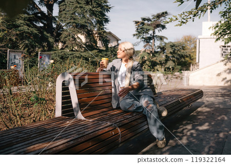 A blonde woman sits on a bench with a cup of coffee in her hand. She is wearing a gray jacket and jeans. The bench is wooden and has a curved back. The scene is peaceful and relaxing. A blonde woman sits on a bench with a cup of coffee in her hand. She is wearing a gray jacket and jeans. The bench is wooden and has a curved back. The scene is peaceful and relaxing. 119322164