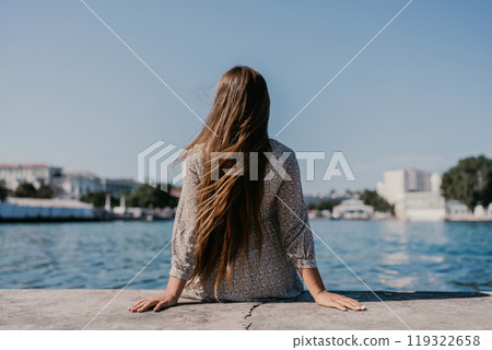 A woman is sitting on a ledge by a body of water, wearing a dress with a leopard print pattern. The scene is peaceful and serene, with the woman enjoying the view of the water and the surrounding area 119322658