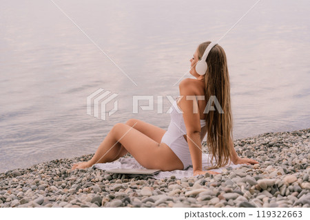 A woman is sitting on a beach with her headphones on. She is wearing a white bikini and a white shirt. The beach is rocky and the water is calm. The woman is enjoying her time by the water. A woman is sitting on a beach with her headphones on. She is wearing a white bikini and a white shirt. The beach is rocky and the water is calm. The woman is enjoying her time by the water. 119322663