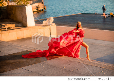 Sunrise red dress. A woman in a long red dress against the backdrop of sunrise, bright golden light of the sun's rays. The concept of femininity, harmony. 119322818