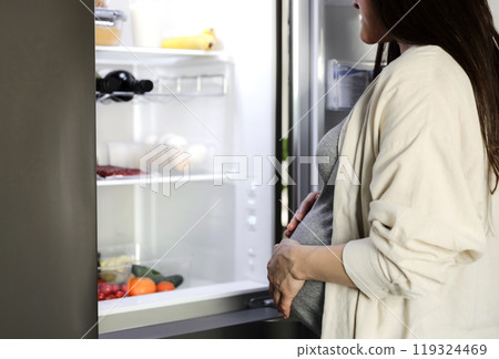 Thoughtful hungry pregnant woman standing in front of open fridge, refrigerator in middle of night at home in kitchen, closeup. Healthy maternity eating, nutrition, pregnancy diet. Looking for food 119324469